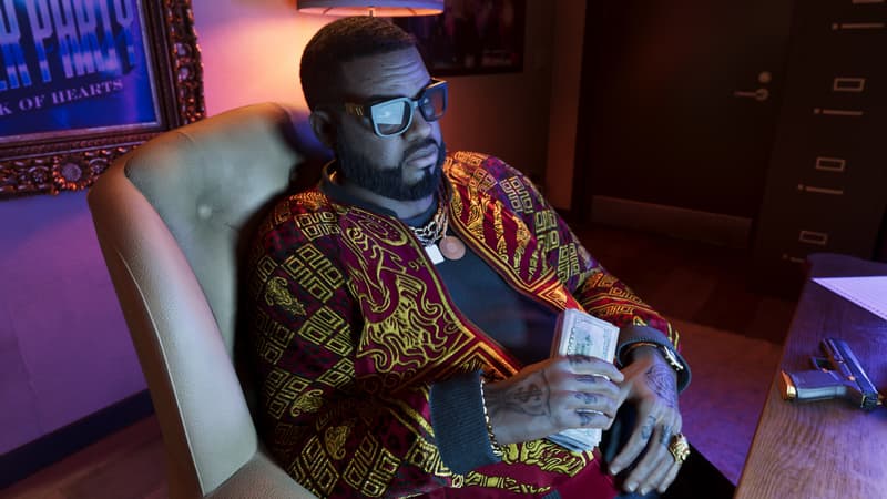 A bearded man in a gold-patterned jacket sits back in an armchair holding a wad of cash, pistol on the desk.