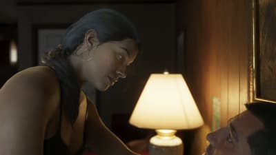 Lucia leans over Jason beside a lit table lamp in a dim motel room at night