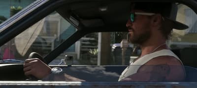 Jason riding shotgun past pastel Keys bungalows, a pink car pulling out ahead