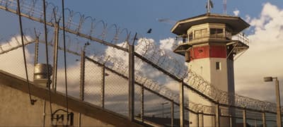 Prison watchtower rising above razor wire and chain-link under a bright blue sky
