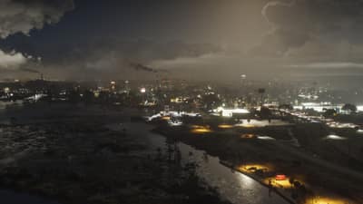 Night aerial over a low industrial town, smokestacks trailing smoke above dark flooded marsh.