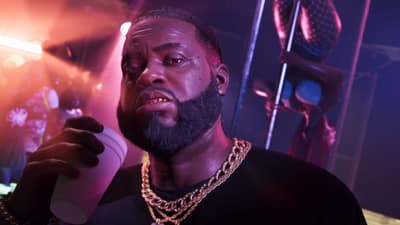 A bearded man in gold chains holds a foam cup inside a pink-lit club.