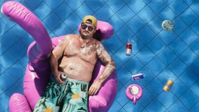 Overhead view of a man in a cap and sunglasses adrift on a pink flamingo pool float.