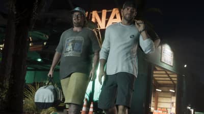 Two men walk away from a lit marina workshop at night, one carrying a white duffel bag.