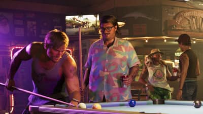 A man in a tank top lines up a shot at a bar pool table under neon signs.