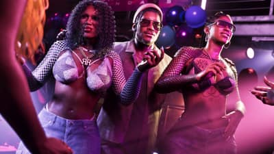 A man in a cream blazer and gold sunglasses poses between two dancers under club lights.