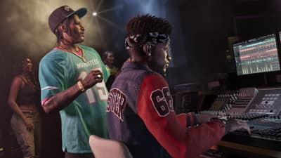 A man in a teal football jersey watches a producer in a bandana work a studio mixing desk.