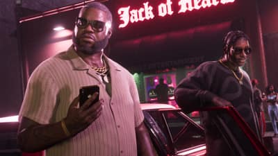Two men in gold chains stand outside the neon Jack of Hearts club sign at night.