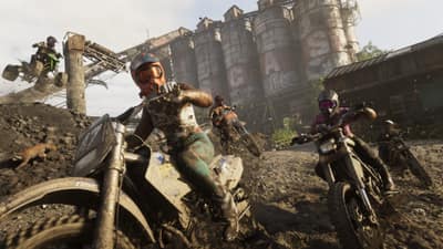 Riders race dirt bikes through mud past derelict grain silos, a wild boar running alongside.