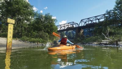 A kayaker paddles a green river toward a steel truss bridge past a yellow flood gauge.