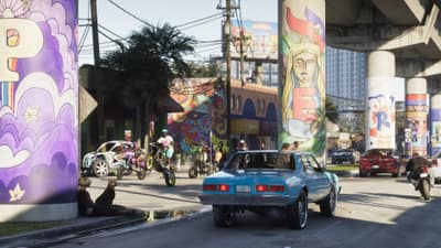 A blue classic sedan and dirt bikes on a street under mural-painted expressway columns.