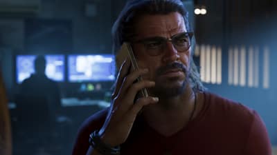 A grey-haired man in glasses takes a phone call in a dim room lined with monitors.