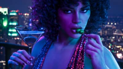 A woman in a sequinned halter eats an olive beside a martini, city lights far below.