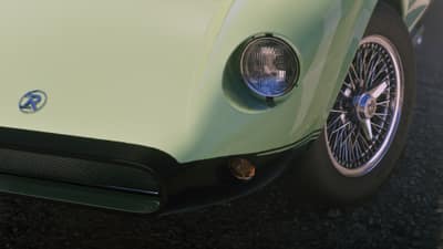 The front corner of a pale green sports car with a chrome wire wheel and round headlight.