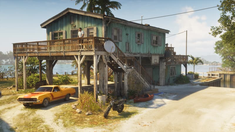 A weathered beach house on stilts with an orange coupe utility, a dirt bike and a kayak parked on the sand.