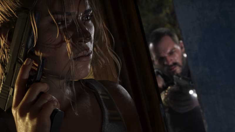 A woman holds a black polymer 9mm pistol upright against her chest in a dim room, an armed man behind her.