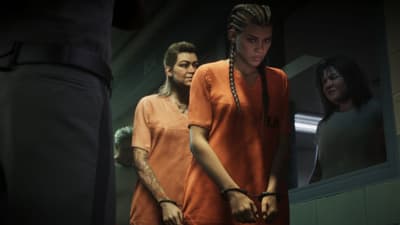 Handcuffed women in orange jumpsuits file past a uniformed guard in a prison corridor.