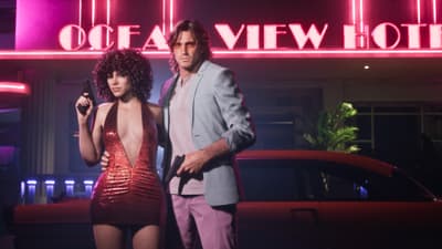 A man and woman in eighties clothes hold pistols on the sidewalk beneath the Ocean View Hotel's pink neon sign.