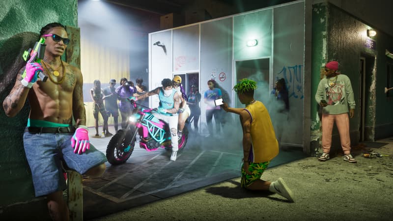 A man with a pink and green pistol leans on a barrier while a rider revs a matching dirt bike outside a store.