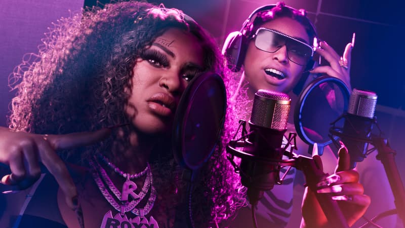 Two women rap into studio microphones under purple light, one in headphones and rhinestone shades.