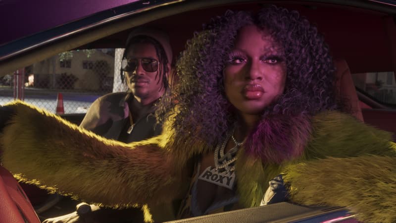 A woman in a yellow fur coat and a diamond Roxy nameplate rides in a car's passenger seat at dusk.
