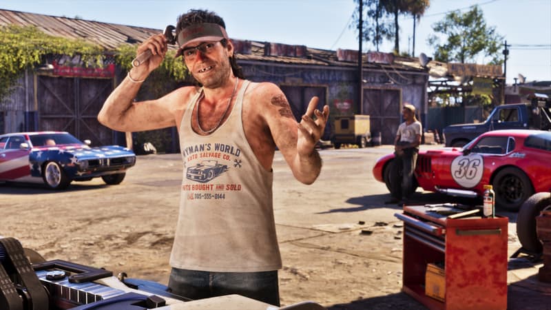 A grinning man in a visor and Wyman's World Auto Salvage vest holds a wrench in a yard of classic cars.